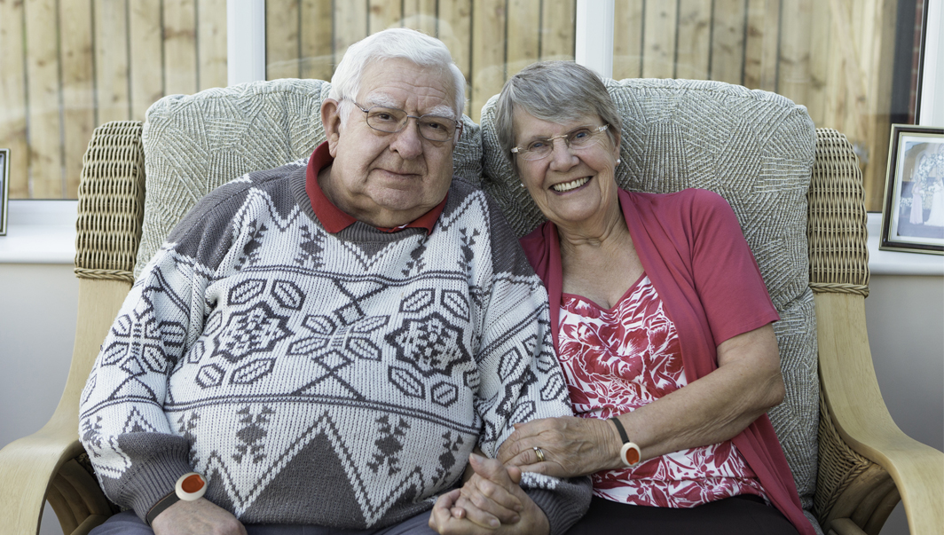 A mature couple sit together hand in hand on the sofa in their conservatory. They are a real couple and looked relaxed and content together.